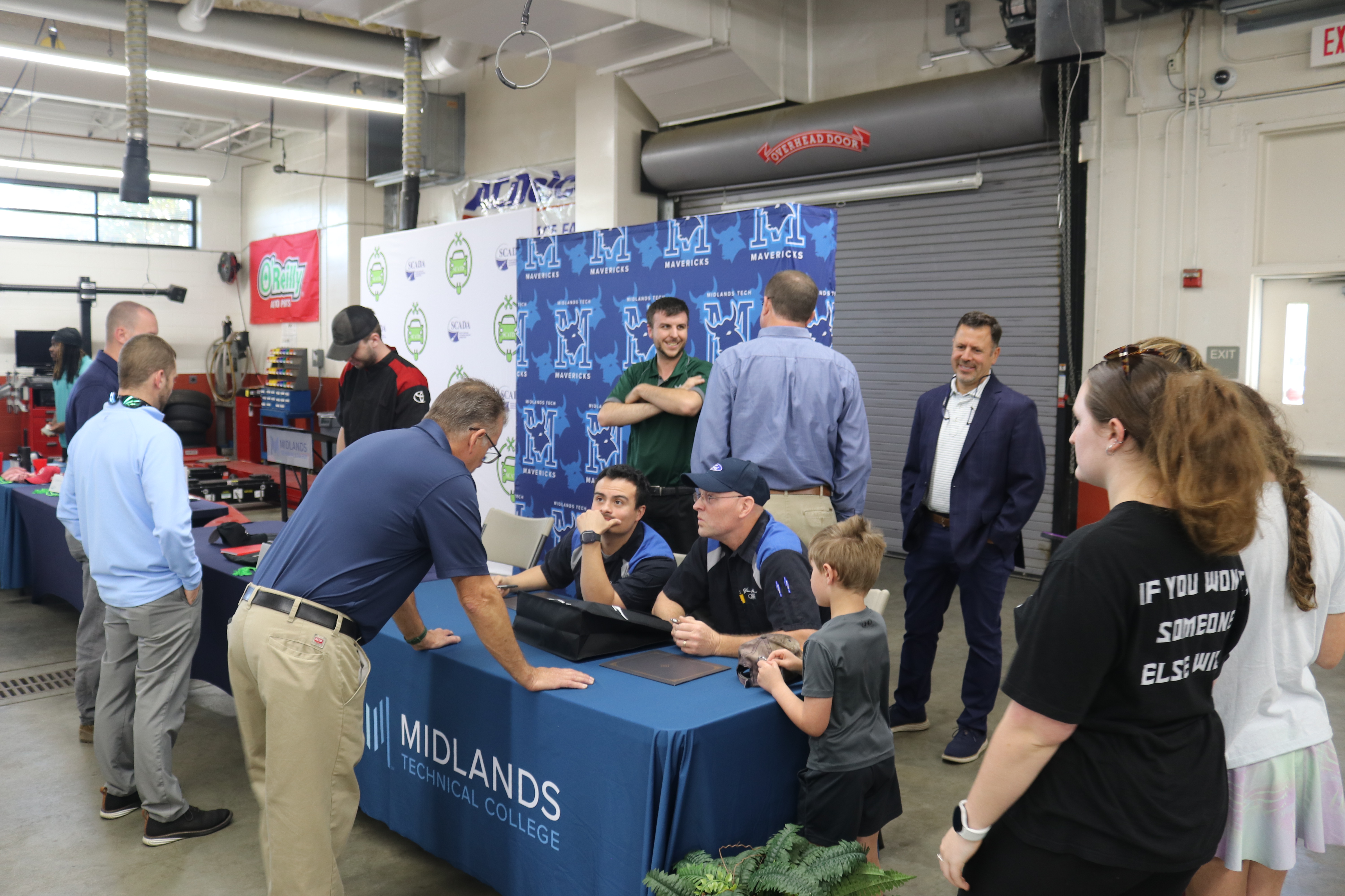 A group of people gather around two apprentices sitting at a table. The people include dealership representatives and family members of the apprentice.