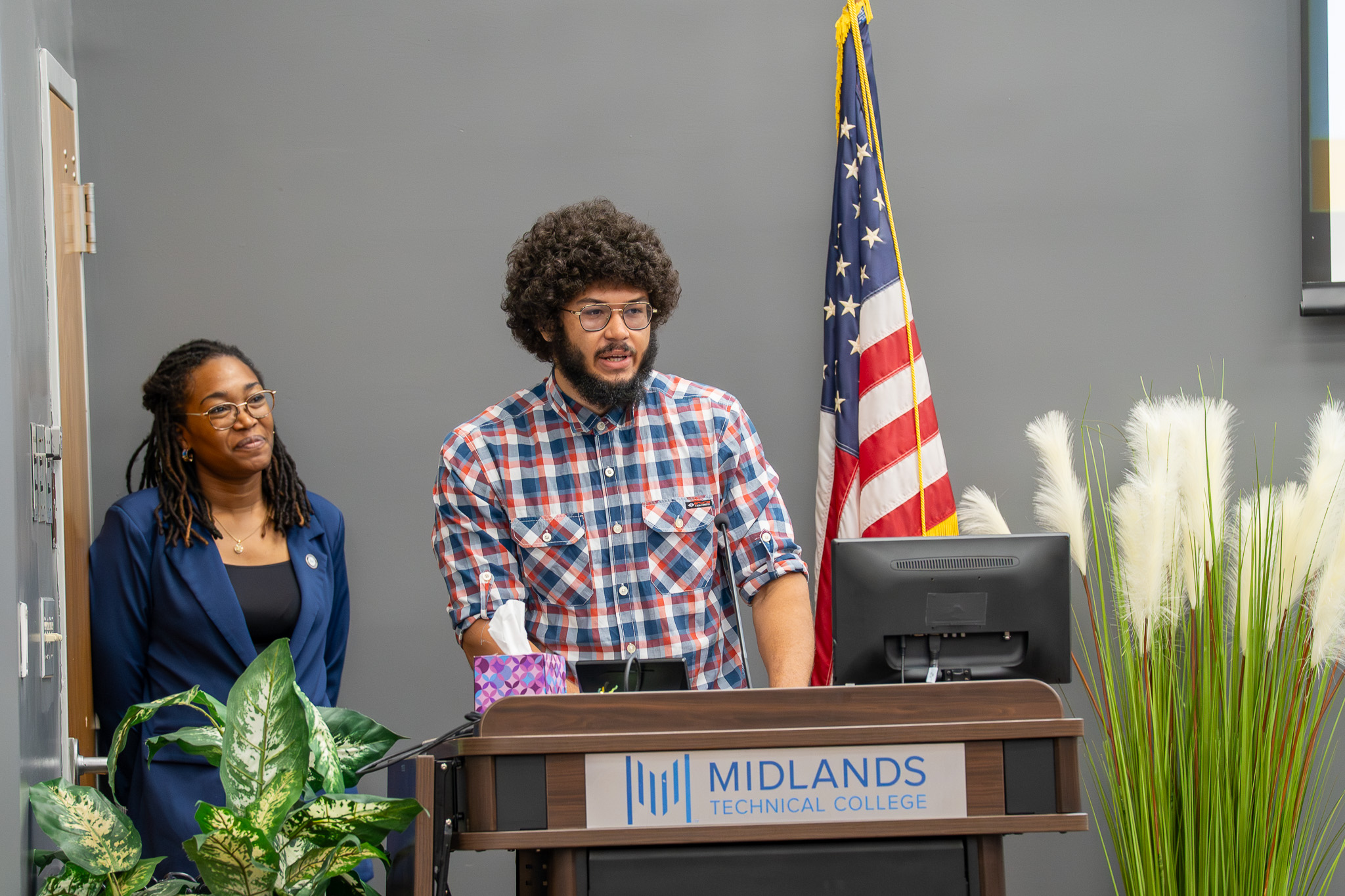 Program Director, Rose-Lynn Heath stands smiling as BOOM graduate, Ryan Robinson delivers the keynote speech.