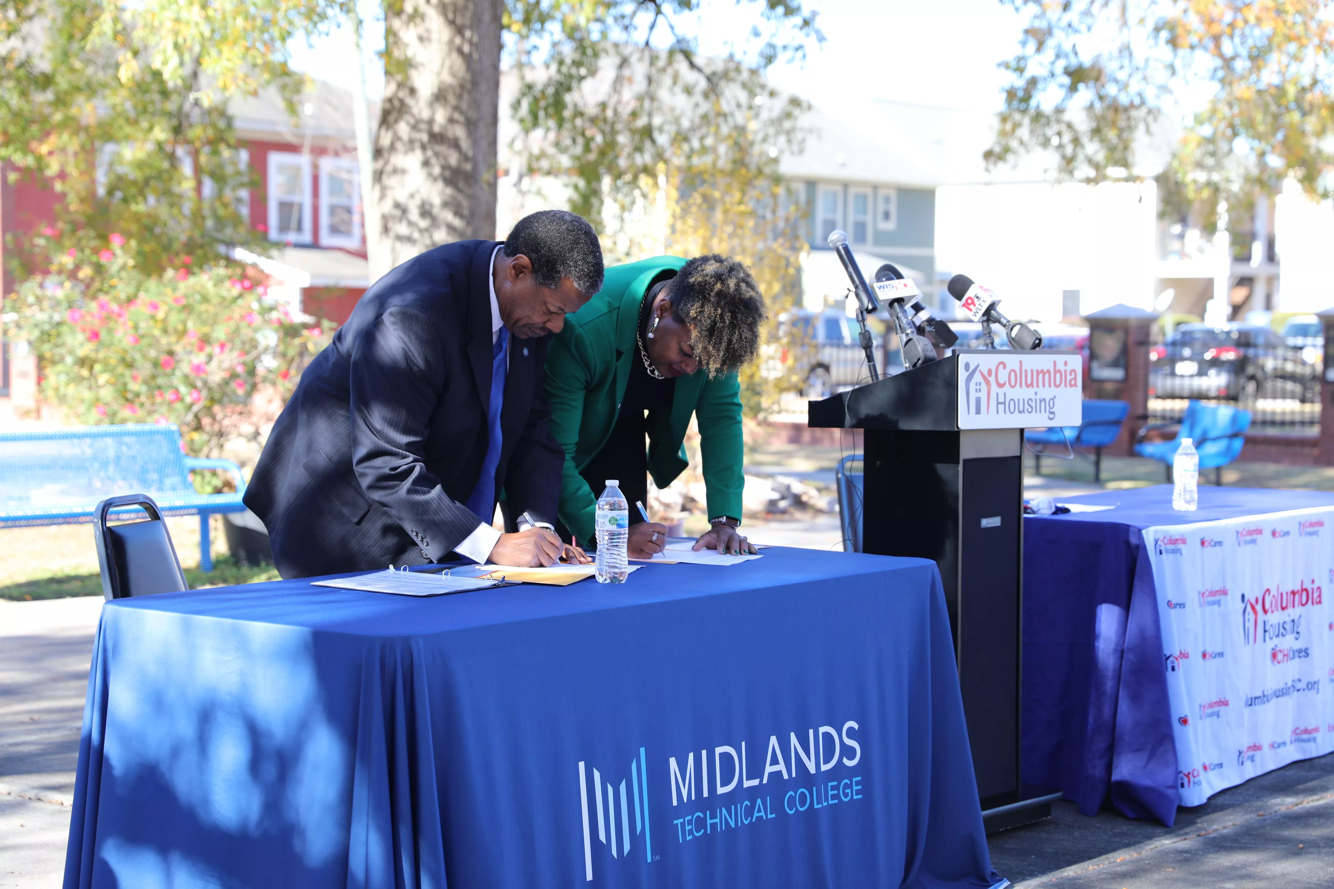 A man and woman signing documents at an outdoor press conference